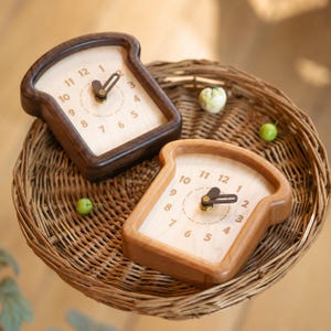 Op de afbeelding: Twee analoge klokken in de vorm van sneetjes brood, één gemaakt van donker hout en de andere van licht hout. Elke klok heeft een lichtgekleurde wijzerplaat met gedrukte cijfers van 1 tot 12 en donkerbruine wijzers, geplaatst op een mand.