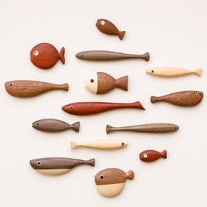 Puede incluir: Una colección de trece figuras de peces de madera tallada en varias formas y tamaños. Los peces están hechos de diferentes tonos de madera, que van desde el beige claro hasta el marrón medio y rojizo, cada uno con un pequeño ojo circular.