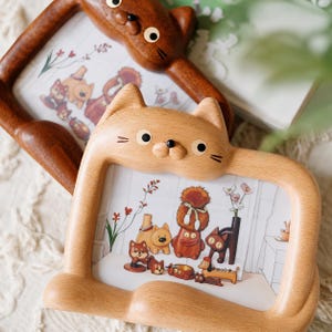 May include: Two rectangular wooden picture frames shaped like cats. The front frame is light-toned wood, while the rear frame is a darker brown wood. Both frames feature carved cat ears, whiskers, and a nose, and contain an illustration of various small animal figurines.