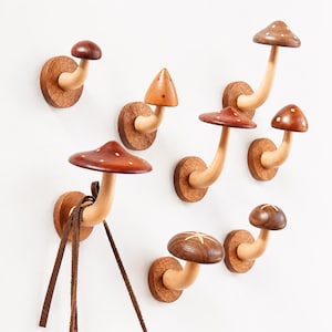 Peut inclure: Un ensemble de sept patères murales en forme de champignons en bois. Chaque patère se compose d'une base circulaire, d'une tige courbée et d'un chapeau dans des tons bruns variés, certains ornés de points blancs ou d'étoiles gravées.