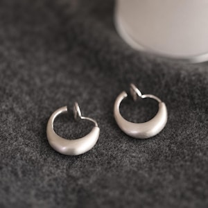 Op de afbeelding: Een paar zilverkleurige oorringen met een gladde, matte afwerking. De oorbellen hebben een halvemaanvormig ontwerp dat naar boven toe taps toeloopt, waar een klein, dun draadje zichtbaar is om de ring op het oorlelletje vast te zetten.
