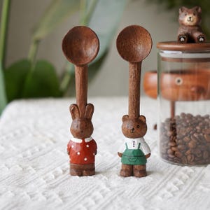 Peut inclure: Deux cuillères en bois sculpté avec des manches en forme d'animaux. Une cuillère a un manche en forme de lapin marron portant une chemise rouge à pois blancs. L'autre a un manche en forme d'ours marron portant une chemise blanche et un tablier vert.