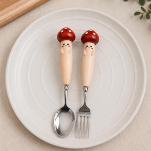 Può includere: Un set di cucchiaio e forchetta in acciaio inossidabile con manici in legno chiaro a forma di fungo. Ogni manico presenta un cappello rosso con pois bianchi, un volto dipinto con piccoli occhi e bocca, e due minuscole braccia laterali.
