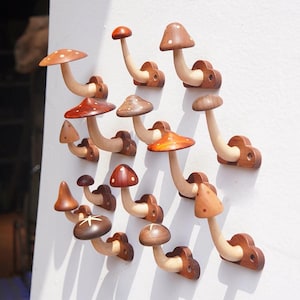 May include: A collection of fourteen wall-mounted mushroom-shaped hooks crafted from various shades of wood. Each hook features a curved stem and a cap, with some caps displaying light-colored spots or star-shaped inlays, mounted on small wooden bases.