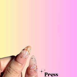 Puede incluir: Primer plano de dos u&ntilde;as postizas con dise&ntilde;os intrincados. Una u&ntilde;a presenta un dise&ntilde;o de concha con detalles dorados, mientras que la otra tiene un dise&ntilde;o brillante con pedrer&iacute;a. Las u&ntilde;as est&aacute;n sobre un fondo degradado rosa y amarillo con el texto "Press To Impress" y "We made the best handmade nails for you!"