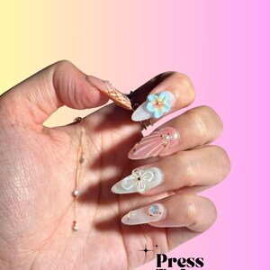 Puede incluir: Un conjunto de u&ntilde;as postizas largas y blancas con una variedad de dise&ntilde;os. Algunas u&ntilde;as tienen un acabado nacarado, mientras que otras tienen un dise&ntilde;o floral o un acento de purpurina dorada. Las u&ntilde;as se muestran en una mano con una delicada pulsera de cadena dorada.
