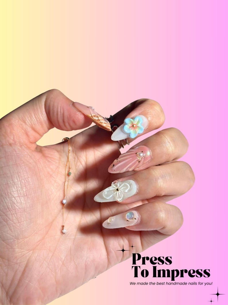 Puede incluir: Un conjunto de u&ntilde;as postizas largas y blancas con una variedad de dise&ntilde;os. Algunas u&ntilde;as tienen un acabado nacarado, mientras que otras tienen un dise&ntilde;o floral o un acento de purpurina dorada. Las u&ntilde;as se muestran en una mano con una delicada pulsera de cadena dorada.