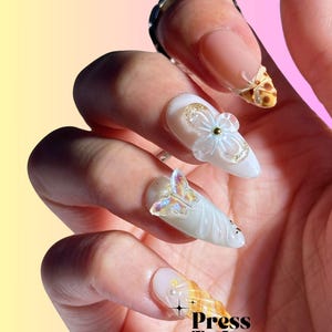 Puede incluir: Primer plano de una mano con u&ntilde;as postizas blancas y azul claro decoradas con flores 3D, mariposas y remolinos. Las u&ntilde;as est&aacute;n decoradas con purpurina dorada y tienen un acabado brillante. El texto "Press To Impress" es visible en la esquina inferior derecha de la imagen. &iexcl;Hicimos las mejores u&ntilde;as hechas a mano para ti!"