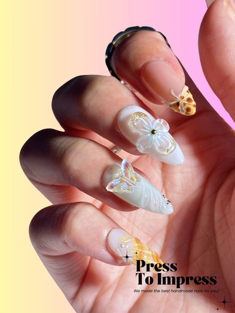 Puede incluir: Primer plano de una mano con u&ntilde;as postizas blancas y azul claro decoradas con flores 3D, mariposas y remolinos. Las u&ntilde;as est&aacute;n decoradas con purpurina dorada y tienen un acabado brillante. El texto "Press To Impress" es visible en la esquina inferior derecha de la imagen. &iexcl;Hicimos las mejores u&ntilde;as hechas a mano para ti!"