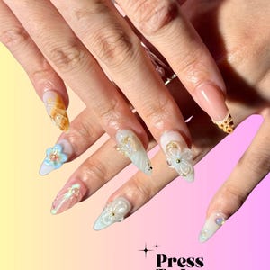 Puede incluir: Primer plano de una mano con u&ntilde;as postizas largas, blancas y azul claro. Las u&ntilde;as est&aacute;n decoradas con flores 3D, remolinos y purpurina. Las u&ntilde;as est&aacute;n sobre un fondo rosa y amarillo con el texto "Press To Impress" y "We made the best handmade nails for you!"