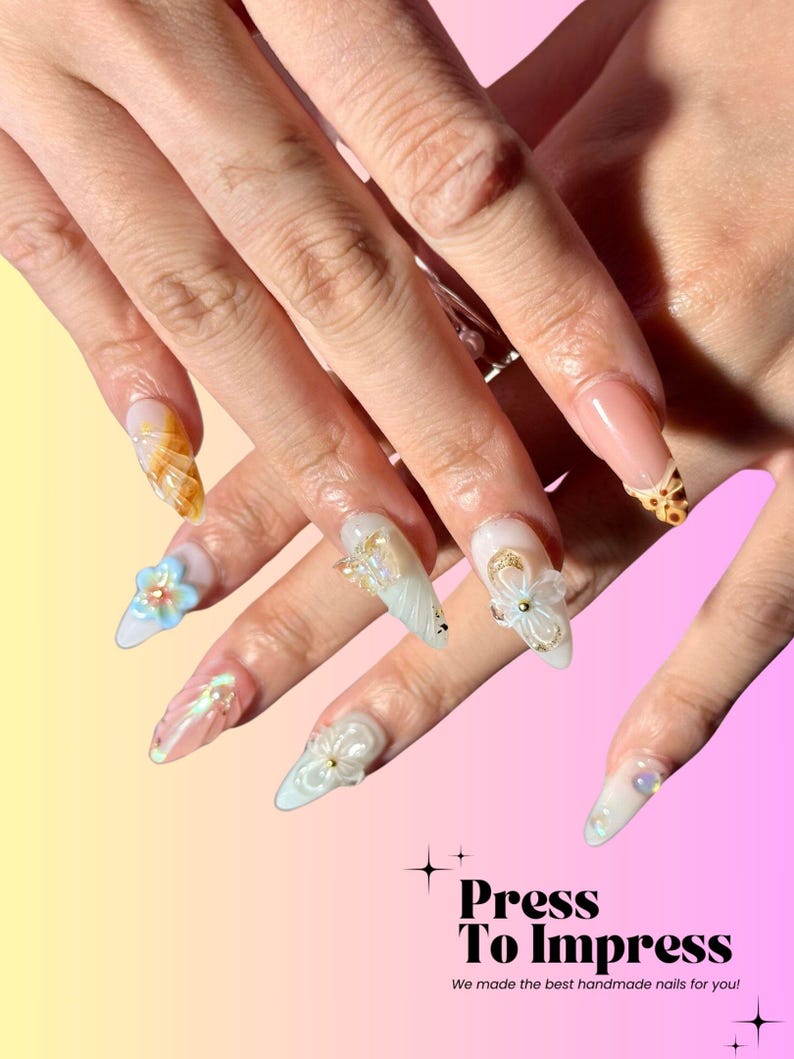 Puede incluir: Primer plano de una mano con u&ntilde;as postizas largas, blancas y azul claro. Las u&ntilde;as est&aacute;n decoradas con flores 3D, remolinos y purpurina. Las u&ntilde;as est&aacute;n sobre un fondo rosa y amarillo con el texto "Press To Impress" y "We made the best handmade nails for you!"