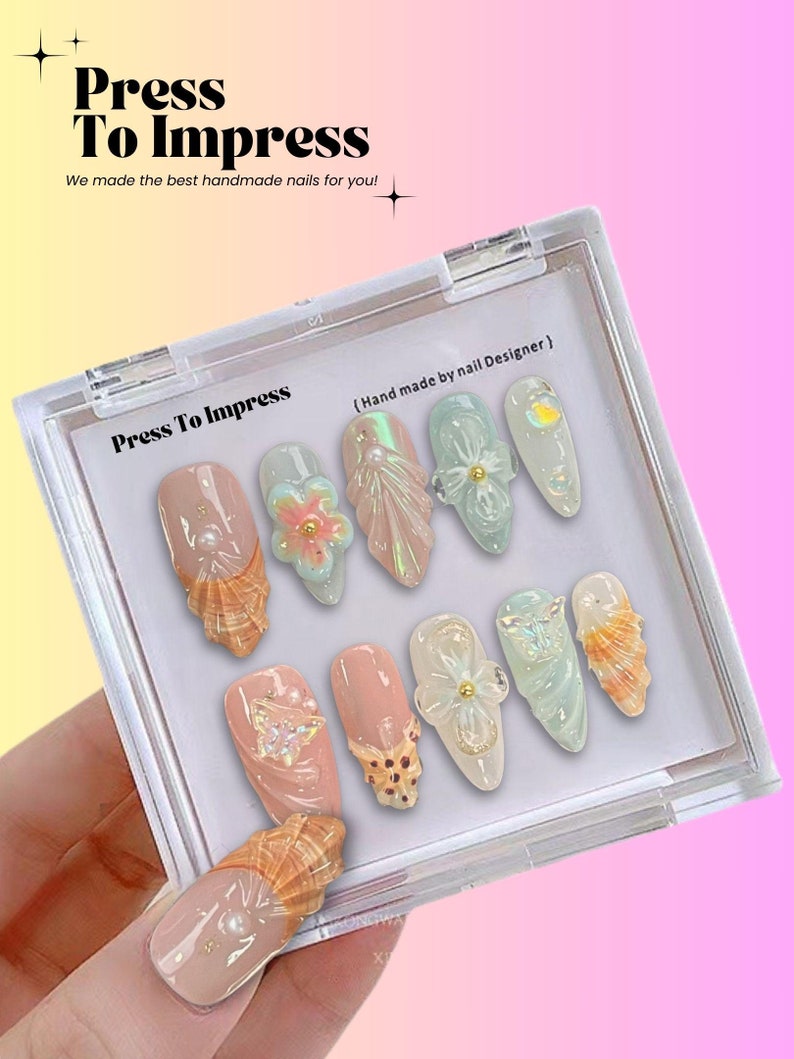 Puede incluir: Un conjunto de diez u&ntilde;as postizas en una caja de pl&aacute;stico transparente. Las u&ntilde;as est&aacute;n decoradas con conchas marinas, flores y purpurina. La caja tiene el texto "Press To Impress" y "(Hand made by nail Designer)" impreso en ella.