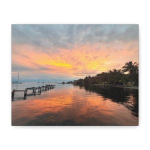 May include: A scenic sunset over a calm body of water with a wooden pier extending into the water. The sky is a vibrant orange and pink, reflecting in the water. Palm trees line the shore.