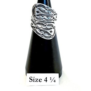 May include: A silver bypass ring featuring intricate stamped geometric patterns and oxidized detailing. The ring is displayed on a black cone stand with a white label at the base clearly printed with the text Size 4 1/4, indicating the ring size.