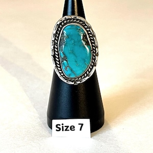 May include: A silver ring featuring a large, oval-shaped turquoise stone with dark matrix veining. The stone is set in a decorative silver bezel with a twisted rope border and etched detailing along the edges. A white tag labeled Size 7 is attached.
