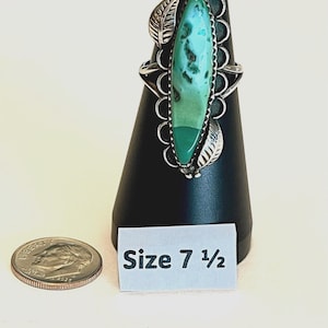 May include: A silver ring featuring a long, marquise-shaped turquoise stone with a two-tone green color variation. The setting includes decorative silver leaf accents and scalloped edges. A small white sign displays the text Size 7 1/2.