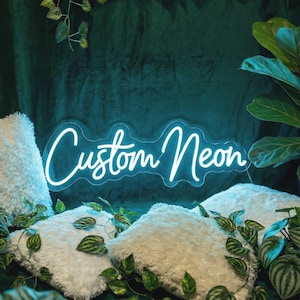 May include: A bright blue neon sign featuring the text Custom Neon in a cursive script font. The sign is mounted on a clear acrylic backing and emits a vibrant glow, positioned among white fluffy pillows and green leafy plants.