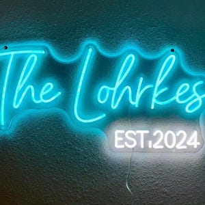 May include: A neon sign in a teal color displays the words "The Lohrkes" in a cursive font. Below, in a white rectangular box, the text "EST. 2024" is displayed. The sign is mounted on a textured teal wall.
