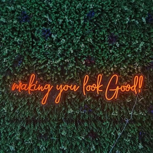 May include: An orange neon sign with the words "making you look Good!" is mounted on a wall of green foliage. The sign's cursive font and bright color contrast with the natural background, creating a visually appealing display.