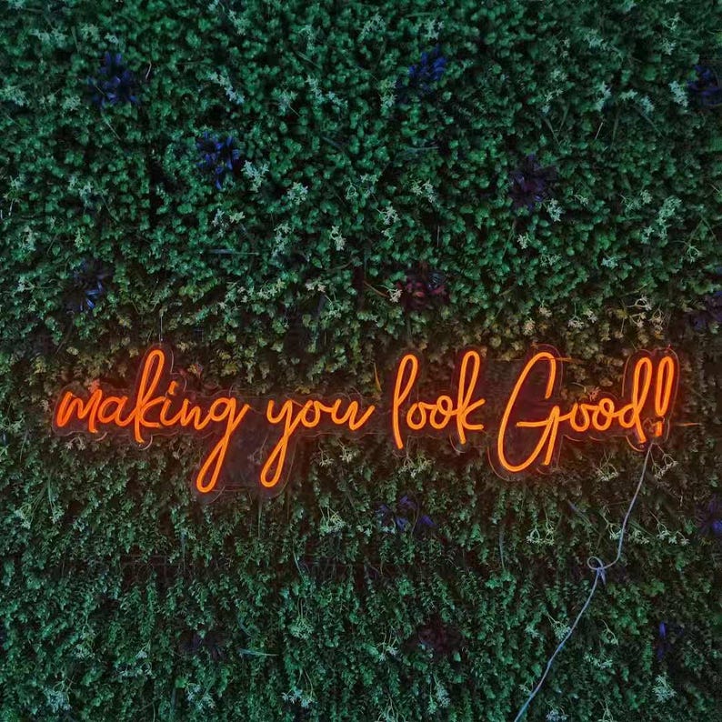 May include: An orange neon sign with the words "making you look Good!" is mounted on a wall of green foliage. The sign's cursive font and bright color contrast with the natural background, creating a visually appealing display.