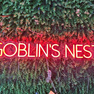 May include: A neon sign with the words "GOBLIN'S NEST" in a bright yellow font, outlined in red. The sign is mounted on a wall of green foliage with small pink flowers.