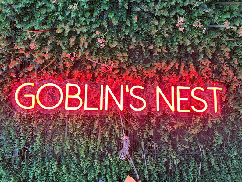 May include: A neon sign with the words "GOBLIN'S NEST" in a bright yellow font, outlined in red. The sign is mounted on a wall of green foliage with small pink flowers.