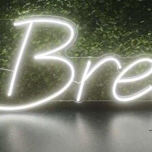 May include: A white neon sign displaying the text "The Breauxs". The sign is illuminated against a backdrop of green foliage. The neon lights are bright and the letters are in a cursive font.