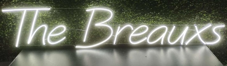 May include: A white neon sign displaying the text "The Breauxs". The sign is illuminated against a backdrop of green foliage. The neon lights are bright and the letters are in a cursive font.