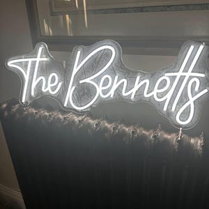 May include: A white neon sign that reads "The Bennetts" in a cursive font. The sign is mounted on a dark, textured surface, possibly a radiator. The sign is illuminated, creating a bright contrast against the dark background.