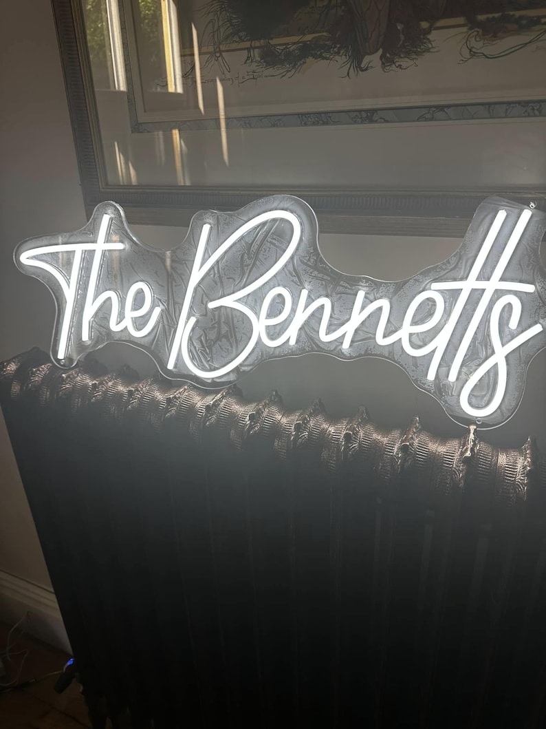 May include: A white neon sign that reads "The Bennetts" in a cursive font. The sign is mounted on a dark, textured surface, possibly a radiator. The sign is illuminated, creating a bright contrast against the dark background.