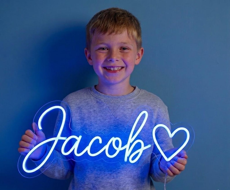 May include: A neon sign in the shape of the name "Jacob" in a cursive font, with a heart symbol. The sign glows with a bright blue light. The sign is held by a smiling child against a blue background.