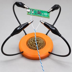 Op de afbeelding: Een groen Raspberry Pi Pico microcontroller-bord vastgehouden door vier zwarte krokodillenklemmen op flexibele armen, geplaatst boven een oranje ronde basis met een messing draad soldeerpuntreiniger en twee witte draden.