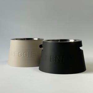 Peut inclure: Deux gamelles pour animaux avec des inserts en acier inoxydable. L'une est beige avec le nom REGGIE en relief sur le côté, et l'autre est noire avec le nom LENNY en relief sur le côté. Les deux ont une base large et évasée avec une poignée.