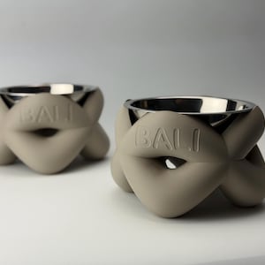 Könnte beinhalten: Zwei Futternäpfe mit glänzendem silberfarbenem Metallinnenraum und einer matten beigen Außenseite in skulpturalem, verknotetem Design. Das Wort BALI ist auf der Vorderseite jedes Napfes eingeprägt. Die Näpfe haben eine abgerundete Form.