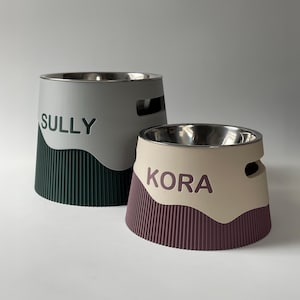 Pode incluir: Dois comedouros para animais com inserções de aço inoxidável. O mais alto é cinza com uma base canelada verde-escura e o nome SULLY. O mais baixo é bege com uma base canelada bordô e o nome KORA. Ambos possuem aberturas laterais.