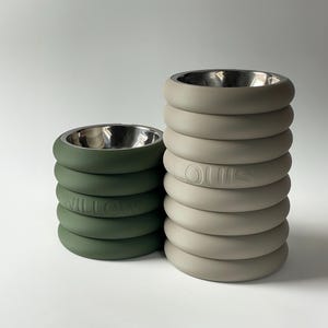 Op de afbeelding: Twee cilindrische voerbakken met metalen binnenkant en gestapelde, afgeronde buitenringen. De kortere bak is donkergroen met de naam WILLOW in reliëf. De hogere bak is lichtbeige met de naam LOUIS in reliëf op de zijkant aangebracht.