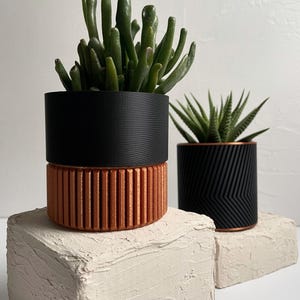 Puede incluir: Dos macetas de plantas negras y cobre con suculentas. Las macetas tienen un diseño texturizado y están sobre bloques blancos texturizados.