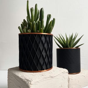 Puede incluir: Dos macetas cilíndricas negras con bordes de cobre, cada una con una suculenta diferente. La maceta más grande tiene un patrón de diamantes, mientras que la más pequeña presenta un diseño de chevrón. Ambas están sobre una superficie texturizada blanquecina.