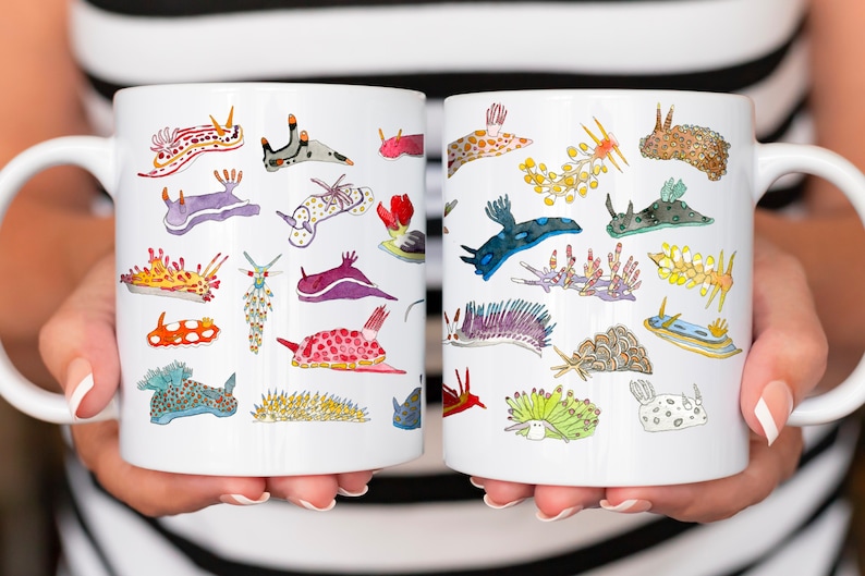 May include: Two white ceramic mugs with colorful illustrations of various sea slugs. The mugs are held by hands and the handles are facing outward.