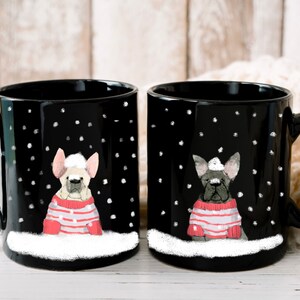 May include: Two black ceramic mugs with white snowflakes and a white border. Each mug features a cartoon French bulldog wearing a red and white striped sweater and a white Santa hat. The mugs are on a white wooden surface.