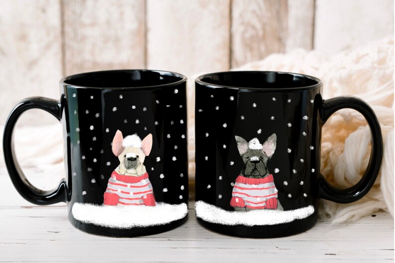 May include: Two black ceramic mugs with white snowflakes and a white border. Each mug features a cartoon French bulldog wearing a red and white striped sweater and a white Santa hat. The mugs are on a white wooden surface.