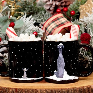 May include: Two black mugs with white polka dots and winter scenes. Each mug has a dog illustration and a candy cane sticking out of the top. The mugs are filled with marshmallows and are surrounded by Christmas decorations.