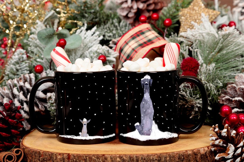 May include: Two black mugs with white polka dots and winter scenes. Each mug has a dog illustration and a candy cane sticking out of the top. The mugs are filled with marshmallows and are surrounded by Christmas decorations.