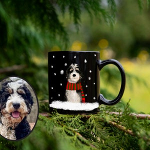 May include: Black ceramic mug with a white dog wearing a red and green scarf in a snowy scene. The dog is a doodle breed with black and white fur.