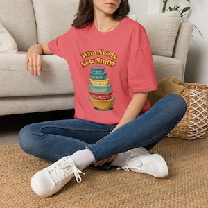 May include: A coral-colored short-sleeve t-shirt featuring a graphic of a stack of six colorful mixing bowls. Above the bowls, the text reads "Who Needs New Stuff?" in a curved, brown, retro-style font. The bowls display various patterns.