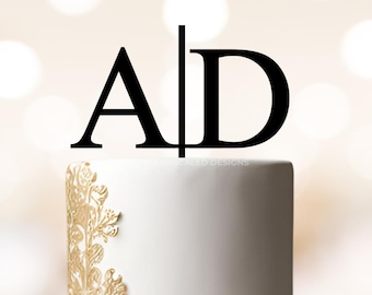 May include: A two-tiered white wedding cake with gold floral accents and a black acrylic cake topper with the letters "A" and "D" separated by a vertical line.