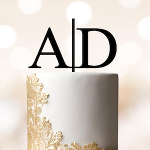 May include: A two-tiered white wedding cake with gold floral accents and a black acrylic cake topper with the letters "A" and "D" separated by a vertical line.