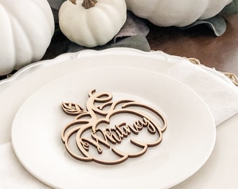 May include: A laser-cut wooden pumpkin-shaped place card featuring the name Whitney in a cursive script font. The brown wooden cutout is centred on a white ceramic plate, which is part of a layered table setting with a white napkin.