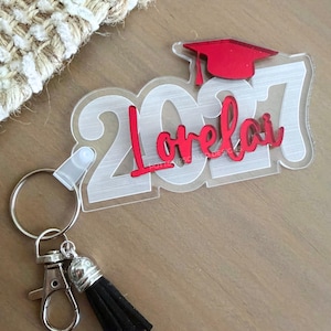 May include: A clear acrylic keychain featuring the white numbers 2027, a red graduation cap icon, and the name Lorelai in red script lettering. The keychain includes a silver metal split ring, a lobster clasp, and a black faux suede tassel.