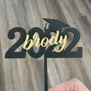 Pu&ograve; includere: Decorazione per torta di laurea nera e oro con l'anno 2022 e il nome "brody". La decorazione include una silhouette del cappello di laurea. La decorazione &egrave; tenuta da un sottile bastoncino nero.